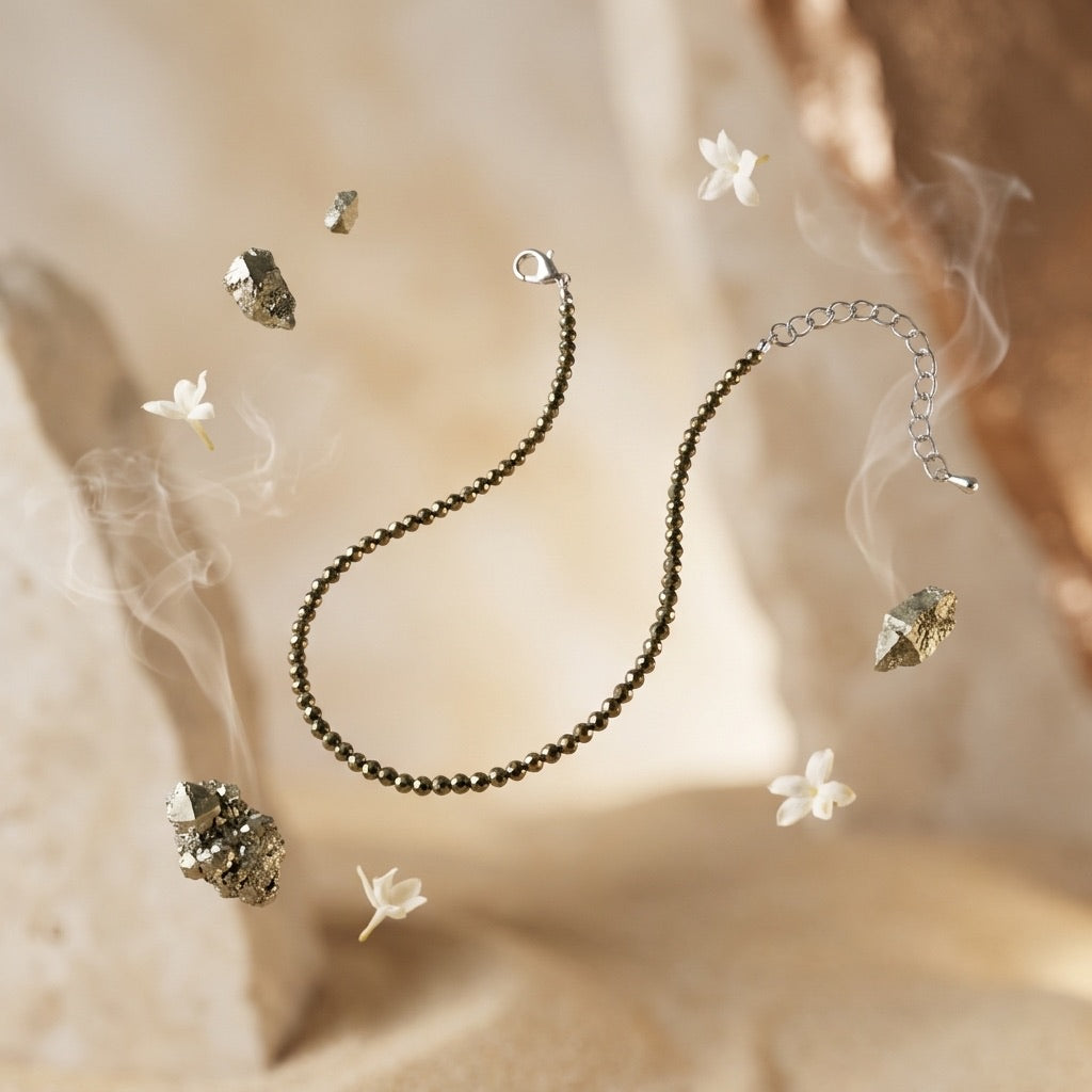 A dark metallic beaded chain anklet with a lobster clasp and extender, styled with crystal clusters and small white flowers against a warm beige textured background.